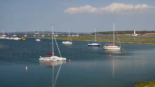 This landscape photograph was taken in the afternoon during late spring and shows several sailing boats and other transport vessels moored on the water at Milford Haven, located in Dorset, United Kingdom. In the distance, Hurst Point Lighthouse is visible on the coastline, providing a recognizable maritime landmark. The calm water reflects the boats, and the shoreline in the background is lined with greenery. The image captures transport on the water, with boats being the central subject against a clear sky and scattered clouds.